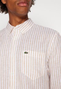 Camicia a righe bianca e beige chiaro realizzata in tessuto testurizzato, con colletto a bottoni e un piccolo logo verde ricamato sulla tasca del petto.