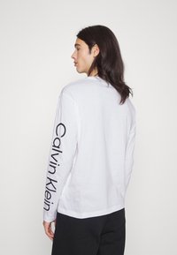Camiseta de manga longa em algodão branco com um logótipo preto "Calvin Klein" na manga esquerda e um design clássico de gola redonda.
