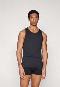Schwarzes Tanktop aus Baumwolle mit runder Ausschnitt und breiten Schulterträgern, kombiniert mit schwarzen Boxershorts, Model steht vor neutralem Hintergrund.