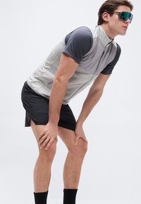 Atletski moški v športnih sončnih očalih, sivi telovnik in črnih kratkih hlačah se nagnil naprej in z rokami naslonil na kolena na enobarvnem ozadju.