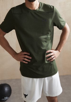 Mann trägt dunkelgrünes kurzärmeliges Hemd und weiße Sportshorts mit Stier-Logo, Hände in die Hüften gestützt, steht neben einem schwarzen Medizinball.
