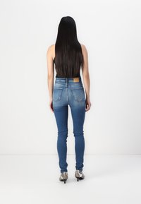 Jeans ajustados de mezclilla azul con una cintura de altura media, bolsillos traseros y una etiqueta de cuero en la cinturilla. El modelo lleva zapatos de tacón plateados.