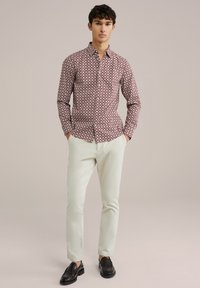 Langsleeve, knoopjesoverhemd met een rood-wit geometrisch patroon. Gepaard met lichtgekleurde broeken en zwarte instappers. Neutrale achtergrond.
