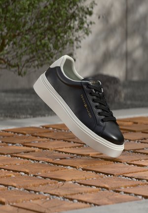 Zwarte leren sneaker met witte zool en hielstuk, zwarte veters en gouden "Tommy Hilfiger"-branding, zwevend boven houten tegels buiten.