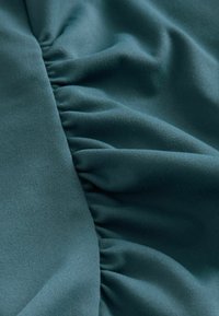 Teal tkanina z gladko teksturo, ki na eni strani prikazuje nabirke, kar ustvarja subtilno globino in dimenzijo.