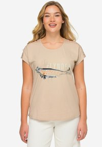 Camiseta de manga corta beige con un estampado gráfico de un pez. Presenta costuras decorativas en los hombros y un corte relajado.