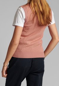 Stickad ärmlös tröja i ljusrosa med strukturerat mönster, ribbad fåll och rundad halsringning, bärs över en vit t-shirt, i kombination med mörka byxor.