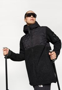 Melna izolētā jaka ar teksturētu flīsa ķermeni un gludu, quilted plecu daļu. Aprīkota ar augstu apkakli un rāvējslēdzēja aizdari, valkāta kopā ar tūrisma nūjām.