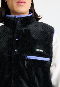 Black fleece vest with a high collar, purple accents, a zippered pocket, and a logo on the chest. Textured, warm fabric with snaps.