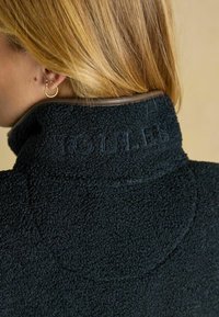 Schwarze Fleecejacke mit hohem Kragen und dem markierten Text "JOULES" in einem strukturierten Stich. Sie verfügt über eine glatte braune Borte am Kragen.
