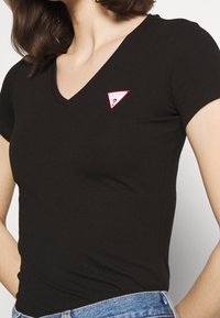 Camiseta negra de manga corta y cuello en V, hecha de una tela suave, con un parche rosa en forma de triángulo del logo "GUESS?" en el lado izquierdo del pecho.