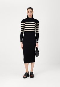 Črna pletena obleka z rebrasto teksturo in kremnimi vodoravnimi črtami, oprijet silhueta, visok ovratnik, dolgi rokavi, v kombinaciji s črnimi platform sandals in torbico.