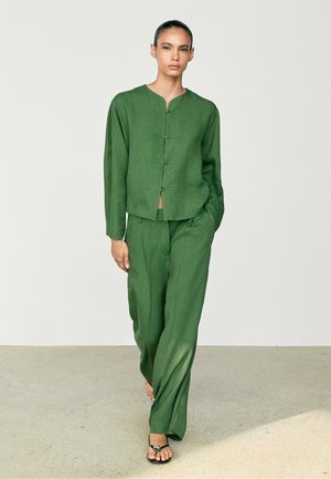 Femme aux cheveux attachés, portant une veste verte boutonnée et un pantalon large assorti, marchant pieds nus sur un sol clair.