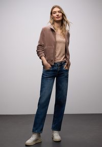 Nainen seisoo kädet taskuissa, yllään vaaleanlila vetoketjullinen takki, eläinkuosi toppi, siniset farkut ja valkoiset lenkkarit harmaalla lattialla.