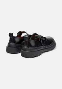 Een paar zwarte leren Mary Jane schoenen met dikke rubberen zolen en verstelbare gespbandjes, weergegeven van achteren op een witte achtergrond.