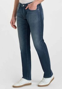 Jean en denim bleu foncé avec une coupe droite, cinq poches et un léger délavage. Associé à des baskets blanches avec des semelles beiges.