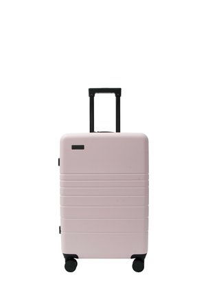Valise rigide rose clair avec poignée noire télescopique et quatre roulettes noires, debout sur fond blanc.