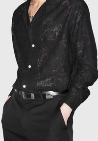 Camicia nera in lino con una texture leggera e semi-trasparente, davanti con bottoni e colletto, abbinata a pantaloni neri su misura e una cintura argentata.