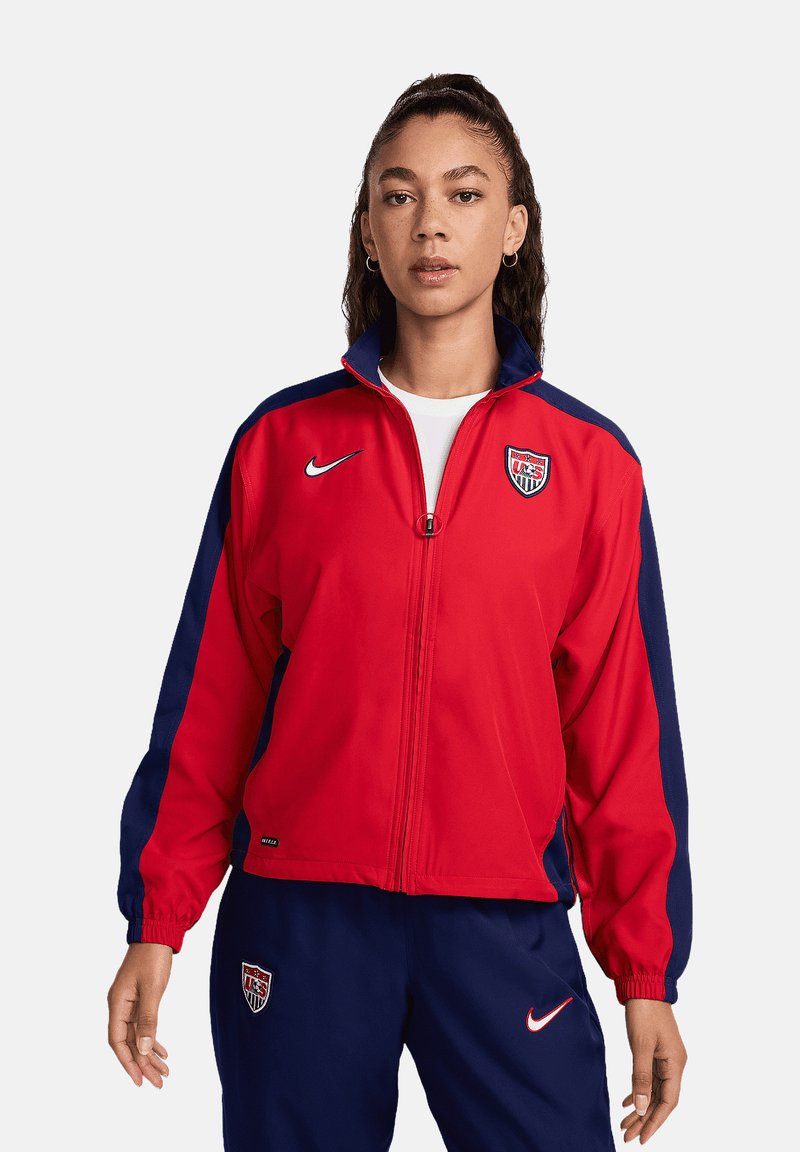 Veste zippée rouge avec des manches bleu marine, ornée d'un logo Nike et d'un emblème de l'équipe nationale des États-Unis sur la poitrine. Tissu lisse avec une coupe sportive.