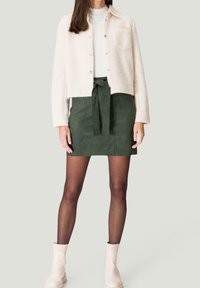 Mujer con una chaqueta corta crema, top blanco, falda mini verde de ante con cinturón de lazo, medias negras semitransparentes y botas cortas crema, de pie.