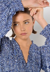 Blaue Bluse mit Muster, V-Ausschnitt und Rüschenkanten. Mit weiten, poetischen Ärmeln. Der Stoff hat ein leichtes, strukturiertes Aussehen.