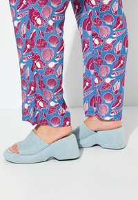 Modré kalhoty s vzorem, na kterých jsou mušle a mořské motivy, doplněné světle modrými klínovými sandály s jemnou texturou látky.