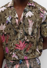 Braune kurzärmlige Hemdbluse mit pinken, roten und lila Blüten- und Insektenmotiven, getragen in passenden gemusterten Hosen.