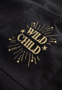 Złoty haftowany tekst "WILD CHILD" z dekoracyjnymi liniami i gwiazdkami na czarnej kieszeni z tkaniny.