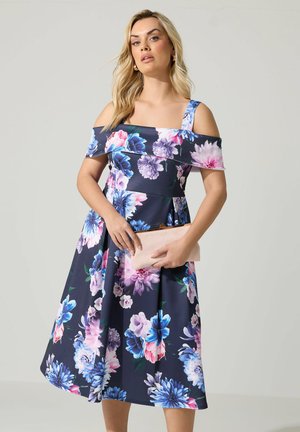 Kvinde iført en marineblå off-shoulder kjole med pink, lilla og blå blomsterprint, der holder en lyserød clutch.