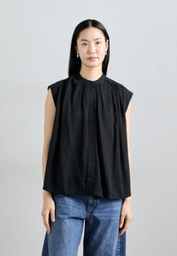 Blouse sans manches noire avec un col haut, des détails plissés et des boutons sur le devant. Associée à un jean en denim bleu clair, avec une coupe décontractée.