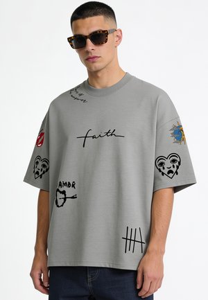 Jonge man met zonnebril met schildpadmotief en een loszittend grijs T-shirt met zwarte grafische prints, waaronder het woord "faith" en hartontwerpen.