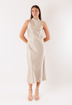 Donna in piedi con le mani dietro la schiena, indossa un vestito midi senza maniche in satin beige e sandali beige con tacchi alti su sfondo bianco.