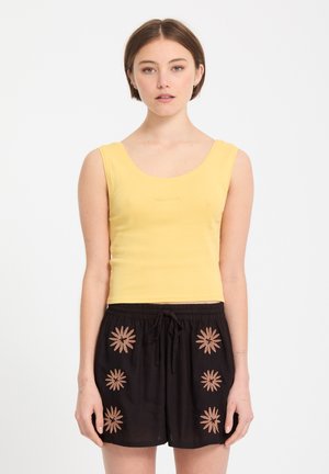 Jeune femme portant un crop top jaune sans manches et un short noir avec des motifs floraux beiges, debout devant un fond blanc.