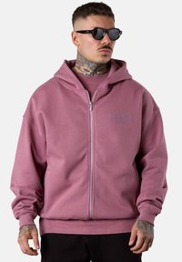 Pink zip-up hoodie with a relaxed fit, kangaroo pockets, ribbed cuffs, and a subtle logo detail on the chest. Smooth cotton blend fabric.