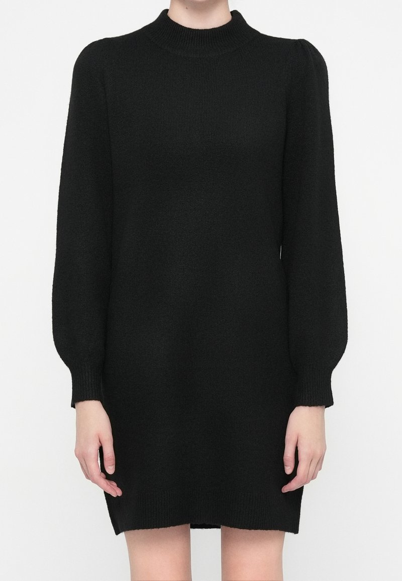 Robe pull noire en maille avec manches longues bouffantes et poignets côtelés, portée par une personne debout devant un fond blanc.
