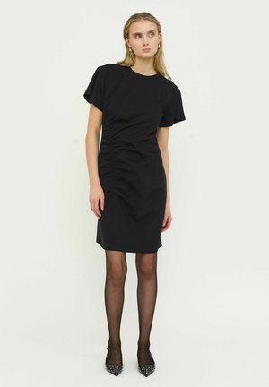 Mujer de pie con un vestido corto negro fruncido de manga corta, medias negras transparentes y tacones puntiagudos negros con tachuelas sobre un fondo liso.