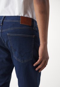 Mörkblå jeans med en läderlogotyp och stickade bakfickor, med en slimmad passform och ren design. Ytan verkar slät.