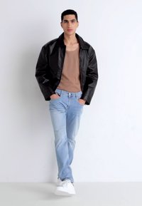Chaqueta de cuero negra sobre una camiseta sin mangas marrón, combinada con jeans azul claro y zapatillas blancas. El modelo está de pie con las manos en los bolsillos frente a un fondo blanco.