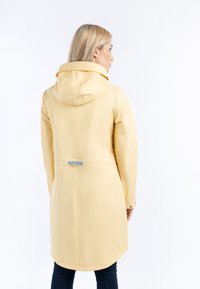 Chubasquero amarillo impermeable con capucha ajustada, cintura ceñida y herrajes con acentos plateados. Textura suave y diseño de longitud media, dobladillo redondeado.