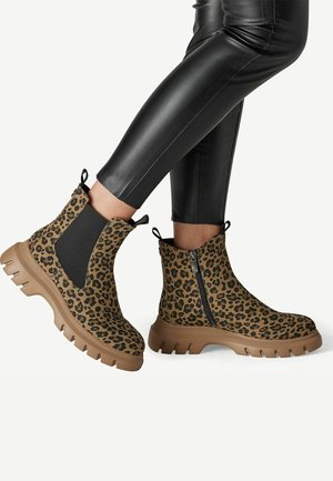 Bottines à imprimé léopard avec semelles épaisses couleur fauve, portées avec des leggings en cuir noirs, montrant une fermeture éclair latérale et des détails de panneaux élastiques.