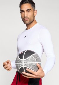 Mann i hvit langermet skjorte som holder en svart-hvitt mønstret basketball, iført røde shorts med svarte snøre.