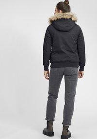 Svart pufferjacka med fuskpäls på huven, ribbade ärmslut och en avslappnad passform, kombinerad med grå jeans och svarta, kraftiga kängor.