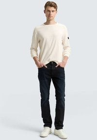 Licht crèmekleurig shirt met lange mouwen, donkere blauwe spijkerbroek en witte sneakers. Het shirt heeft een ronde halslijn en geribbelde manchetten.