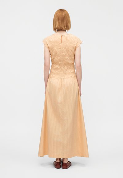 Φόρεμα σε ροδακινί απόχρωση με κοντή φουσκωτή πάνω μέρος και μακριά, αέρινη φούστα με κάθετες ρίγες. Το πίσω μέρος έχει κλείσιμο με κουμπί στον λαιμό.