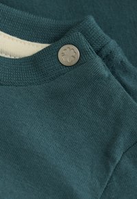 Close-up van teal gebreide stof met een metalen drukknoop, gegraveerd met een klavertje vier, nabij een ribbelrand van de halslijn.