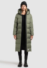 Manteau long et bouffant, couleur vert olive, avec une capuche, doté d'une fermeture éclair noire, d'accents côtelés et de poches latérales ; associé à des bottes noires et un bonnet.