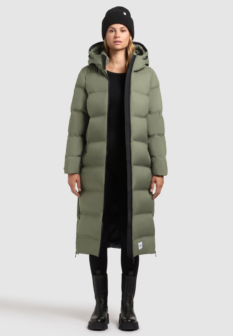 Manteau long et bouffant, couleur vert olive, avec une capuche, doté d'une fermeture éclair noire, d'accents côtelés et de poches latérales ; associé à des bottes noires et un bonnet.