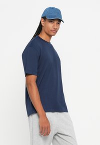 Άτομο με μακριά πλεξούδα που φοράει μπλε καπέλο, ναυτικό μπλε μπλούζα και γκρίζα φόρμα, στέκεται μπροστά σε λευκό φόντο.