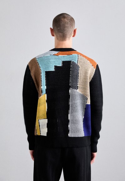 Pull en tricot avec un motif géométrique en noir, bleu, jaune, gris et marron, avec des sections texturées et un col rond.
