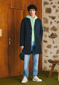 Jūras spēku mētelis virs zaļās kapučas, saskaņots ar gaiši ziliem džinsiem un baltām kedām. Modelis tur melnu cepuri, stāv uz zaļa paklāja pret koka apdari.